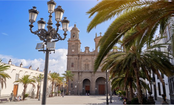 Centro de la ciudad de Las Palmas de Gran Canaria. Imagen tomada desde la Plaza Mayor en la que se encuentra la Basílica de de Santa Ana. La Plaza está rodeada de palmeras y edificios bajos de color blanco. Varias farolas de hierro forjado con cinco lámparas cada una son las encargadas de alumbrar la plaza. Al fono se puede ver la fachada principal de la Basílica de Santa Ana con un alto campanario. 
