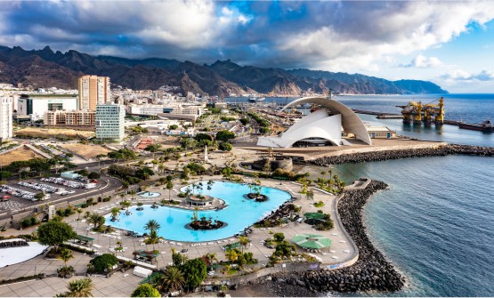 Vista aérea de la ciudad de Santa Cruz de Tenerife con las oscuras montañas al fondo de la imagen y con grandes nubes sobre ellas. En primer plano podemos ver el auditorio, una maravilla arquitectónica vanguardista y todo un símbolo de las Islas Canarias. Frente a él se puede ver la piscina del parque Marítimo César Manrique frente al mar. 