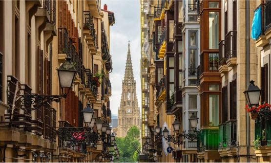 Calle principal del casco antiguo de San Sebastián con la catedral del Buen Pastor al fondo. Una preciosa calle con preciosos edificios repletos de invernaderos adornados con farolas de hierro forjado. Al fondo resalta el campanario de la catedral mientras algunos árboles cubren la base que la sostiene.  