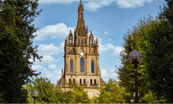 Imagen de la Basílica de Begoña en Bilbao tomada desde el parque donde está ubicada, rodeada por frondosos árboles. En el centro de la imagen surge el campanario de la iglesia de Begoña. En la parte más baja se encuentran varias ventanas desde las que cuelgan muchas campanas. Sobre estas hay tres altas ventanas de medio punto y en el centro un reloj culminando con la cúpula de aguja.  