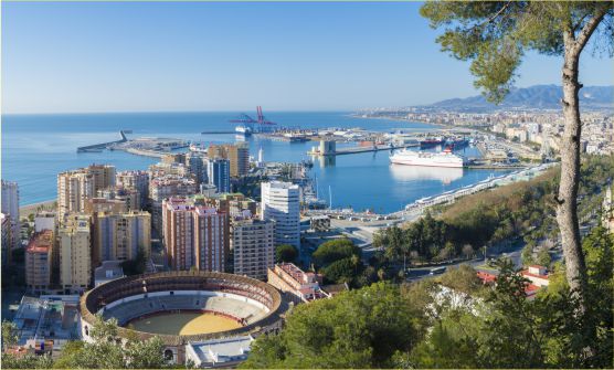 Vista panorámica del puerto de Málaga donde podemos contemplar grandes edificios junto al mar, barcos y una vista aérea de la plaza de toros situada junto a una zona arbolada.