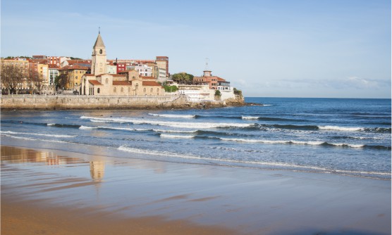 Playa de Gijón desde donde podemos contemplar la iglesia y varios edificios de diferentes colores. Sobre la arena de la playa mojada se refleja la iglesia y el ir y venir de las olas con espuma blanca. 